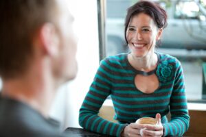 Was passiert, wenn du immer freundlich bist? Eine lächelnde Frau sitzt an einem Tisch gegenüber einem Mann, was eine freundliche und positive zwischenmenschliche Interaktion symbolisiert.